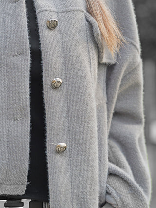 Nahaufnahme einer Person mit der OFF#DLY Jacke Zaira grey, die durch dekorative Metallknöpfe und eine strukturierte Oberfläche besticht, über einem schwarzen Oberteil getragen. Ein Gürtelansatz ist sichtbar.