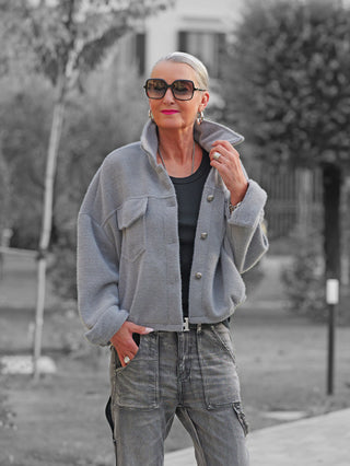 Frau trägt die OFF#DLY Jacke Zaira grey im Park, mit silbernem Haar und Sonnenbrille. Jacke hat markante Knöpfe, Brusttaschen; kombiniert mit schwarzem Top und grauer Cargo-Hose.