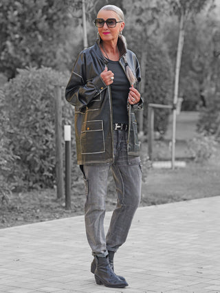 Frau in OFF#DLY Fake Leather Blazer Tatiana black mit markanten Taschen und Oversized-Schnitt, kombiniert mit grauen Cargo-Jeans und schwarzen Stiefeln, steht lässig auf einem Gartenweg.