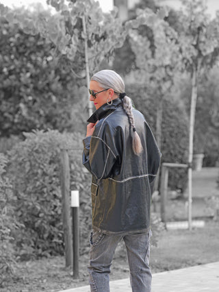 Person im Freien mit hochgezogenem Kragen des OFF#DLY Fake Leather Blazer Tatiana black, oversized geschnitten, in Kombination mit grauen Jeans, langen silbergrauen Zopf und großen Sonnenbrillen.