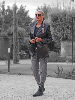 Frau in einem Park trägt den OFF#DLY Blazer Rosalia black, oversized mit Reverskragen und Pattentaschen, kombiniert mit grauen Utility-Hosen und schwarzen bestickten Stiefeletten.