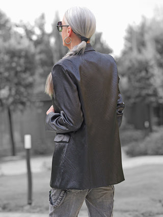 Person von hinten im Park, trägt den OFF#DLY Blazer Rosalia black aus strukturiertem Kunstleder mit Reverskragen und Pattentaschen, kombiniert mit grauen Jeans, große Creolen und Sonnenbrille.