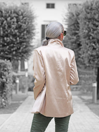 Person von hinten mit dem OFF#DLY Blazer Rosalia champagner in Satin-Optik, mit Reverskragen und Pattentaschen, spaziert auf einem Weg. Der Blazer fällt elegant und strukturiert.