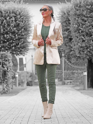 Frau trägt den OFF#DLY Blazer Rosalia champagner mit geradem Schnitt und Reverskragen, kombiniert mit olivgrüner Kleidung, und posiert selbstbewusst vor einem schmiedeeisernen Tor.