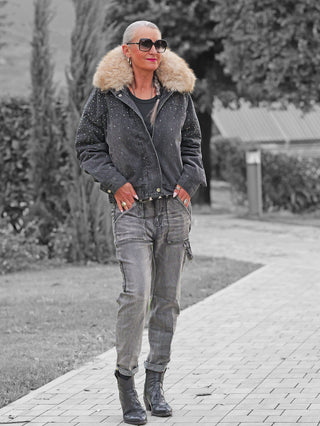 Ältere Dame in OFF#DLY Denim / Fur Jacket Viviana black, mit Kunstfellkragen und Ziersteinen an Ärmeln und Schultern, spaziert auf einem Gartenweg, Hände in den Taschen, trägt Jeans und Lederstiefel.