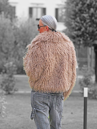 Person von hinten trägt die OFF#DLY Fake Fur Jacke beige aus weichem, voluminösem Faux-Fell, kombiniert mit grauen Cargo-Jeans, im Freien vor unscharfem Hintergrund.