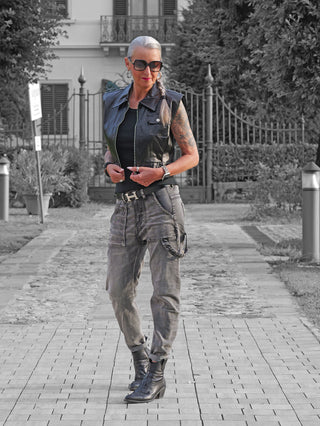 Silberhaarige Frau trägt die OFF#DLY Weste Mariella in Schwarz, kombiniert mit grauen Cargo-Jeans und schwarzen Stiefeletten, vor einem Tor stehend. Die Weste verleiht ihrem Outfit modernen Chic.