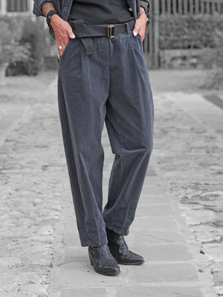 Person trägt OFF#DLY Cord Pant Lavinia anthrazit mit lockerer Passform und passendem Gürtel. Hände in Taschen, schwarze, bestickte Lederstiefel ergänzen den Look auf steinigem Weg.