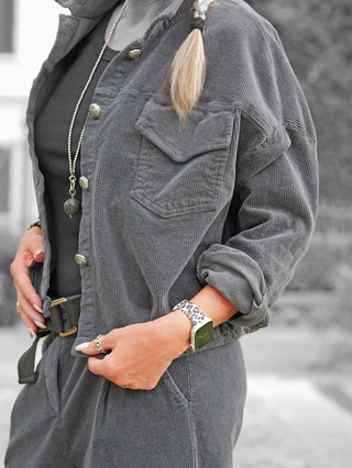 Nahaufnahme einer Person, die die OFF#DLY Cord Jacke Ornella anthrazit trägt, mit aufgerollten Ärmeln, Brusttaschen und dekorativen Knöpfen, kombiniert mit einem schwarzen Top und auffälligen Accessoires.