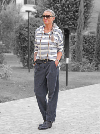 Frau in Kapuzenshirt OFF#DLY LS Kapuze Ingrid white/grey mit Streifen, Kordeln und Kapuze, kombiniert mit lockerer Hose und Stiefeln, steht auf einem gepflasterten Weg.