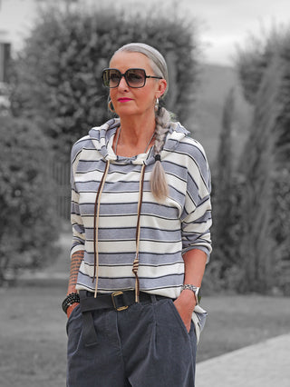 Frau trägt das OFF#DLY LS Kapuze Ingrid white/grey, ein gestreiftes Kapuzenshirt mit langen Kordeln. Sie kombiniert es mit dunklen Cordhosen und Accessoires wie Sonnenbrille und Armbänder.