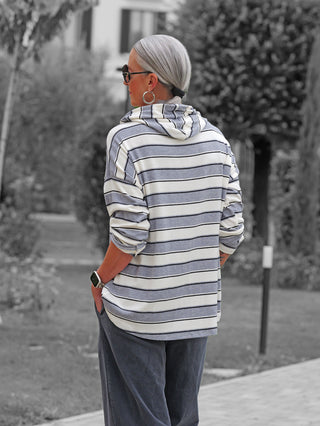 Person von hinten in einem Garten, trägt das OFF#DLY LS Kapuze Ingrid white/grey Kapuzenshirt mit grauen Streifen, lässigem Schnitt und hochgeschobenen Ärmeln, kombiniert mit dunklen Cordhosen.