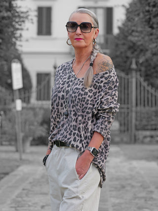 Frau trägt OFF#DLY Longsleeve Astrid beige/black, ein lässig fallendes Oberteil mit Leopardenmuster, über eine Schulter. Kombiniert mit hellen Hosen und auffälligen Accessoires, vermittelt sie Selbstbewusstsein.