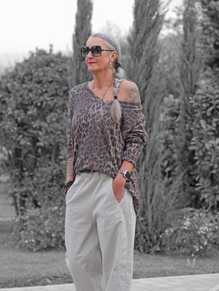 Ältere Frau im OFF#DLY Longsleeve Astrid, einem einseitig schulterfreien Leo-Print-Top mit lockerem Schnitt, kombiniert mit weiten Hosen, Schmuck und Tattoos, in entspannter Pose im Freien.