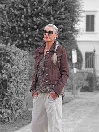 Frau im Freien trägt die OFF#DLY Jacke Bologna schoko, eine schokobraune, fransige Denim-Jacke mit taillierter Form, über einer Leopardenbluse, kombiniert mit hellen, locker sitzenden Hosen.