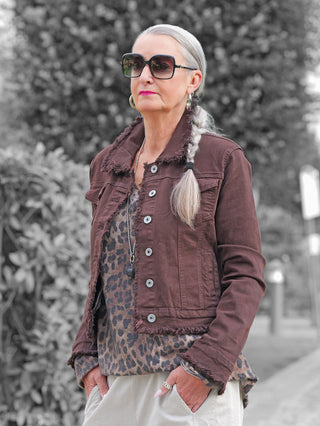 Ältere Frau trägt die OFF#DLY Jacke Bologna schoko mit Fransen, über Leoparden-Top, kombiniert mit hellen Hosen. Die Jacke zeigt eleganten Denim-Stil, passend für Frühling und Sommer.