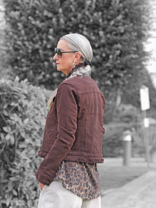 Ältere Frau mit silbernem Zopf trägt die OFF#DLY Jacke Bologna schoko, eine taillierte, leicht gefranste, braune Denim-Jacke, kombiniert mit Leopardenbluse und helle Hosen.