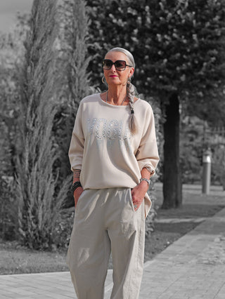 Eine stilvolle Frau trägt das OFF#DLY Sweat Solveig beige mit VOGUE-Schriftzug und funkelnden Applikationen, kombiniert mit weiten Hosen. Der lässige, bequeme Schnitt strahlt schlichte Eleganz aus.
