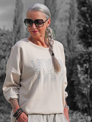 Frau mit silbernem Zopf trägt OFF#DLY Sweat Solveig beige mit VOGUE-Schriftzug in funkelnden Applikationen, kombiniert mit Sonnenbrille und Schmuck, lässig in Taschen der hellen Hose stehend.