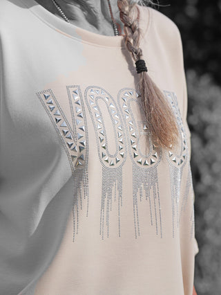 Nahaufnahme des OFF#DLY Sweat Solveig beige: Langarmshirt mit silbernem VOGUE-Schriftzug und pyramidenförmigen Stud-Details, samt Zopf und silberner Kette sichtbar, ideal für glamourösen, lässigen Chic.