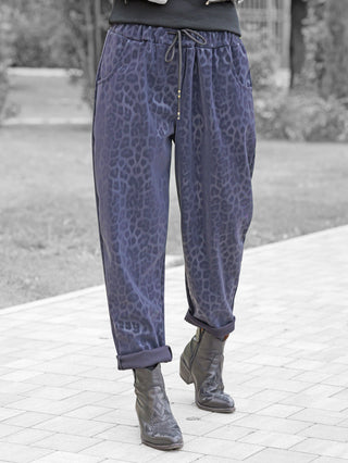 Person trägt die OFF#DLY Leo Pant Freija blue, eine locker sitzende Hose mit dunkelblauem Leopardenmuster und Kordelzug. Kombiniert mit schwarzen Lederstiefeln auf einem gepflasterten Weg.