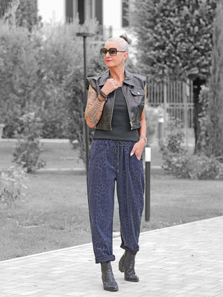 Eine reife Frau trägt die OFF#DLY Leo Pant Freija blue, locker geschnittene Hose mit elastischem Bund und modernem Leopardenmuster, stilvoll kombiniert mit schwarzen Stiefeletten und einem schwarzen Lederweste.
