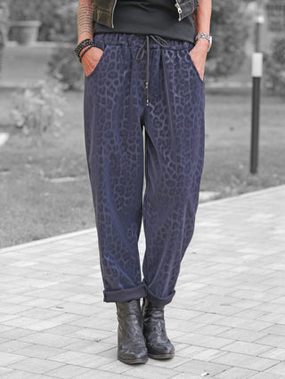Person trägt OFF#DLY Leo Pant Freija blue auf Gehweg, mit lockerem Leo-Print, elastischem Bund und Taschen, kombiniert mit schwarzen Lederstiefeln, schwarzem Top und Lederjacke.
