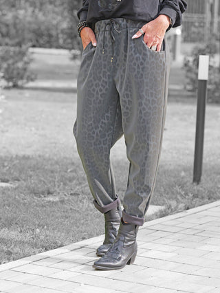 Person trägt OFF#DLY Leo Pant Freija grey mit grauem Leopardenmuster, elastischem Bund und Taschen. Die Hosenbeine sind aufgerollt, kombiniert mit schwarzen Lederstiefeletten. Hände in den Taschen, Schmuck sichtbar.