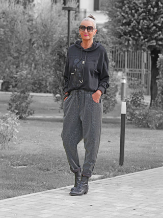 Frau trägt OFF#DLY Leo Pant Freija grey mit lockerem Leo-Print und elastischem Bund. Kombiniert mit schwarzem Hoodie und Boots, steht sie lässig im Park.