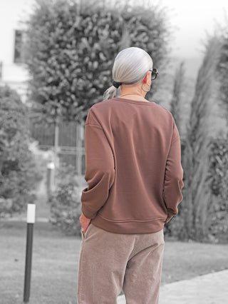 Eine Person trägt den OFF#DLY Sweat Chocolate braun, ein braunes Pullover mit modernem Frontprint, passend zu hellen Kordhosen, während sie auf einem Bürgersteig steht.