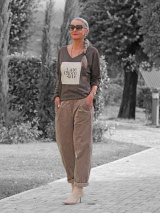 Eine reife Frau mit seitlichem Zopf trägt ein OFF#DLY Longsleeve Latte Choco braun mit Late chocolate-Grafik und entspannter Passform, kombiniert mit hochgeschnittenen Cordhosen und beigen Stiefeletten.
