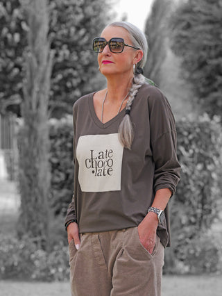 Frau im Freien trägt ein OFF#DLY Longsleeve Latte Choco braun mit entspanntem Schnitt, markantem Frontprint Late chocolate, kombiniert mit hellbraunen Cordhosen und Accessoires wie einer Leopardenarmbanduhr.