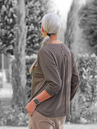 Person von hinten in einem Garten, trägt OFF#DLY Longsleeve Latte Choco braun, lässig geschnitten, silberne Accessoires, kombiniert mit neutralen Cordhosen und einer Smartwatch mit gemustertem Band.