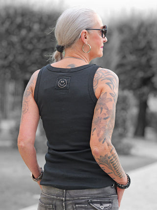 Erwachsene Person von hinten in schwarzem OFF#DLY Top Neve, mit Smiley-Patch, Tattoos, Sonnenbrille. Kombiniert mit Jeans, steht in Garten, tanktop betont lässigen Stil und Baumwollkomfort.