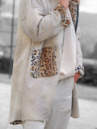 Nahaufnahme des OFF#DLY Mantel Elin stone: Beiger, mittellanger Mantel mit Leopard-Print-Faux-Fur an Kragen und Taschen, über cremefarbenem Oberteil und hellen Hosen, Hand lässig in Seitentasche gesteckt.
