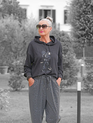 Frau im Garten trägt den OFF#DLY Sweater Pezzei black mit 3D-Teddybär-Motiv und Kapuze, kombiniert mit grauen Leopardenmuster-Hosen und schwarzen Perlenarmbändern.