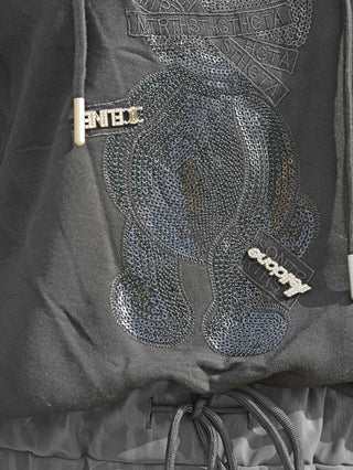 Nahaufnahme des OFF#DLY Sweater Pezzei black: Schwarzer Hoodie mit stilisiertem Teddybär aus schwarzen Pailletten und zwei kleinen Metallanhängern an den Kordeln, die einen edlen Streetstyle-Look verleihen.