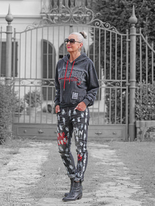 Ältere Frau in OFF#DLY Denim Donatella black/red Jeans mit Graffiti-Optik, kombiniert mit einem grauen Hoodie. Die Jeans hebt sich durch den einzigartigen Streetstyle-Effekt hervor.