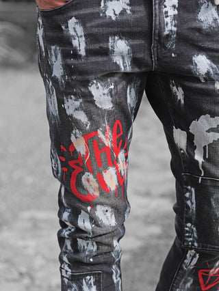 OFF#DLY Denim Donatella black/red Jeans mit weißem Paint-Splatter und rotem Graffiti-Print auf Oberschenkeln; Hand in der Tasche auf Betonhintergrund für Streetstyle-Effekt.