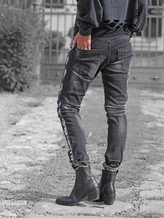 OFF#DLY Denim Donatella black/red Jeans mit helleren Nähten an den äußeren Nähten, schlanke Passform, gekrempelte Knöchel, kombiniert mit schwarzen Lederstiefeletten, auf einem grob gepflasterten Gartenweg stehend.