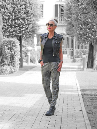 Mature Frau in schwarzem Outfit trägt die OFF#DLY Fake Leather Denim Luna, eine figurbetonte Kunstleder-Hose, kombiniert mit einer ärmellosen Lederweste, verziert mit Schmuck und Tattoos.