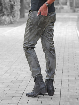 Person trägt OFF#DLY Fake Leather Denim Luna black. Slouchy, lederähnliche Hose kombiniert mit schwarzen Lederstiefeletten. Hand mit Leopard-Uhrenarmband in der Gesäßtasche, Bäume im Hintergrund unscharf.