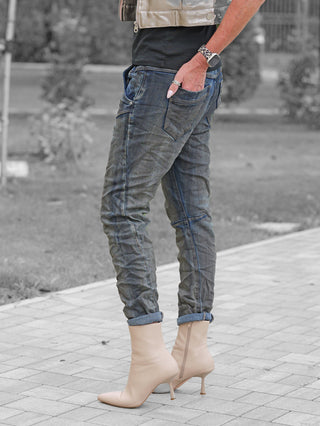 Person trägt OFF#DLY Denim Nika dark blue Jeans mit umgeschlagenem Saum, kombiniert mit spitzen, beigefarbenen Stiefeletten. Eine Hand zeigt ein Leoparden-Armband und einen silbernen Ring.