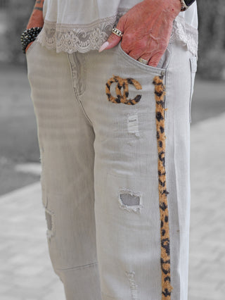 OFF#DLY Denim Mira grey: Hellgraue, distressed Jeans mit schmalem Leoparden-Print-Streifen an der Außenseite, kleinem Leoparden-Print-Emblem nahe der Hüfttasche, kombiniert mit weißer, spitzenbesetzter Bluse und silbernen Ringen.