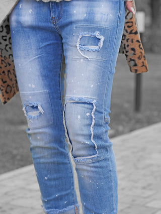 Nahaufnahme der OFF#DLY Denim Lara lightblue mit distressed Rissen an Oberschenkeln und Knien, weißen Farbspritzern und einem Hauch eines Leopardenmantels im Hintergrund.