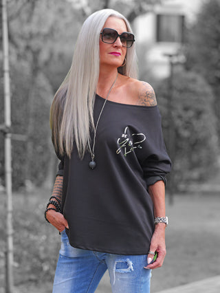 Eine Frau trägt das OFF#DLY Shirt Beauty black, ein schwarzes, schulterfreies Sweatshirt mit schimmerndem Be You tyful-Print, kombiniert mit blauen Jeans, Armbändern und einer Halskette.