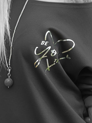 Nahaufnahme des Oberkörpers mit dem OFF#DLY Shirt Beauty black, bedruckt mit BE YOU tiful in Metallic-Folie. Trägt eine Kugelkette mit Lavastein-Anhänger; dezentes Tattoo sichtbar.
