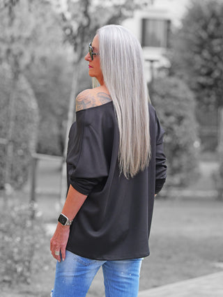 Eine Frau trägt das OFF#DLY Shirt Beauty black, ein lockeres, schulterfreies schwarzes Oberteil mit einem glänzenden Front-Print, kombiniert mit hellen Jeans und einem Tattoo auf der linken Schulter.