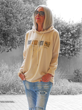 Ältere Frau trägt den OFF#DLY Hoody Alva beige mit Tiermuster-Print und Kapuze, kombiniert mit hellblauen Jeans. Sie steht entspannt mit Händen in den Taschen.