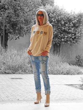 Frau trägt OFF#DLY Hoody Alva beige mit animal-print, kombiniert mit hellen Jeans und beigen Stiefeletten. Hoodie hat lockeren Schnitt und Kapuze, perfekter urbaner Look für Alltag.